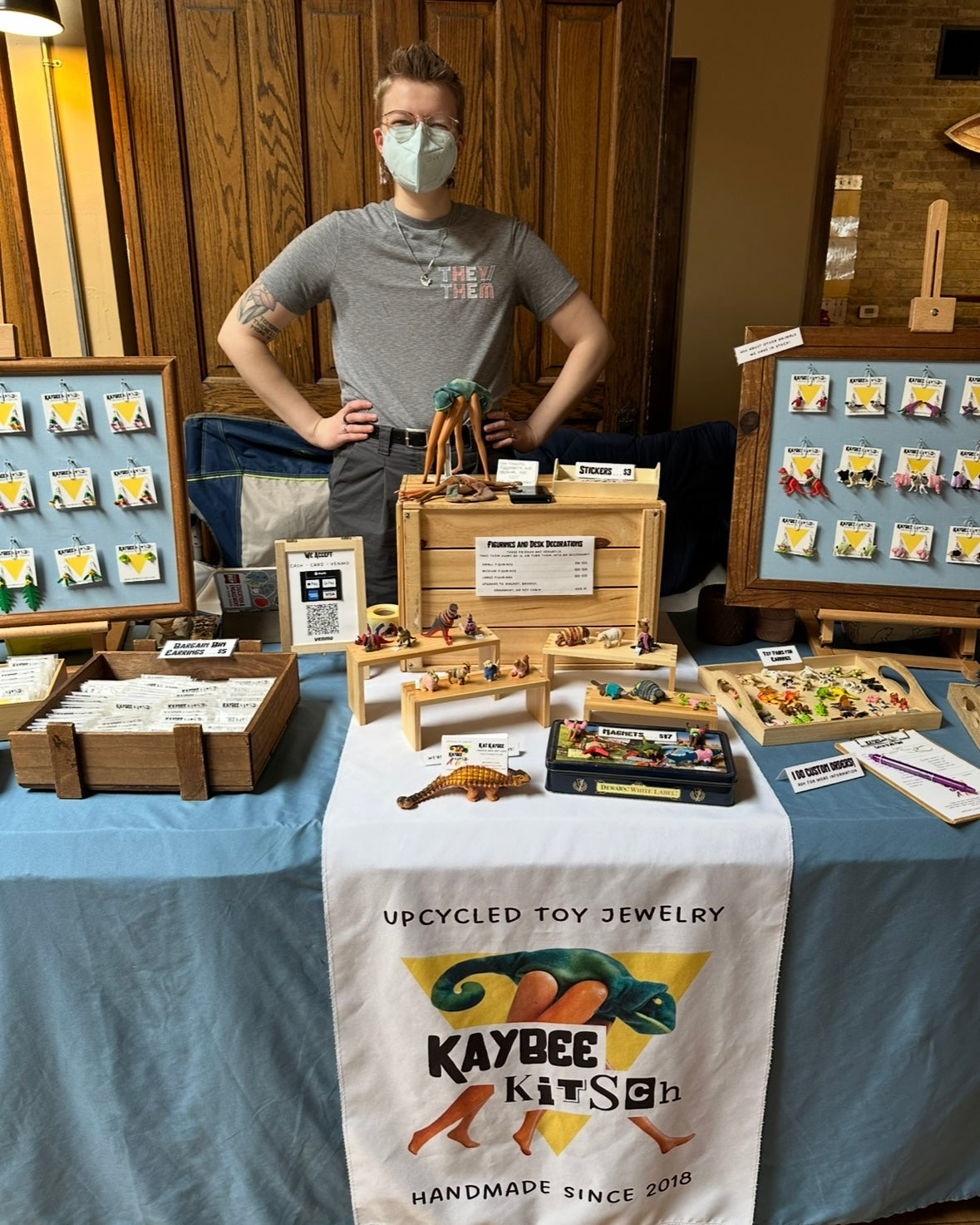 Kat, the business owner, standing behind a table set-up for Kaybee Kitsch displaying earrings, desk figurines, and magnets.