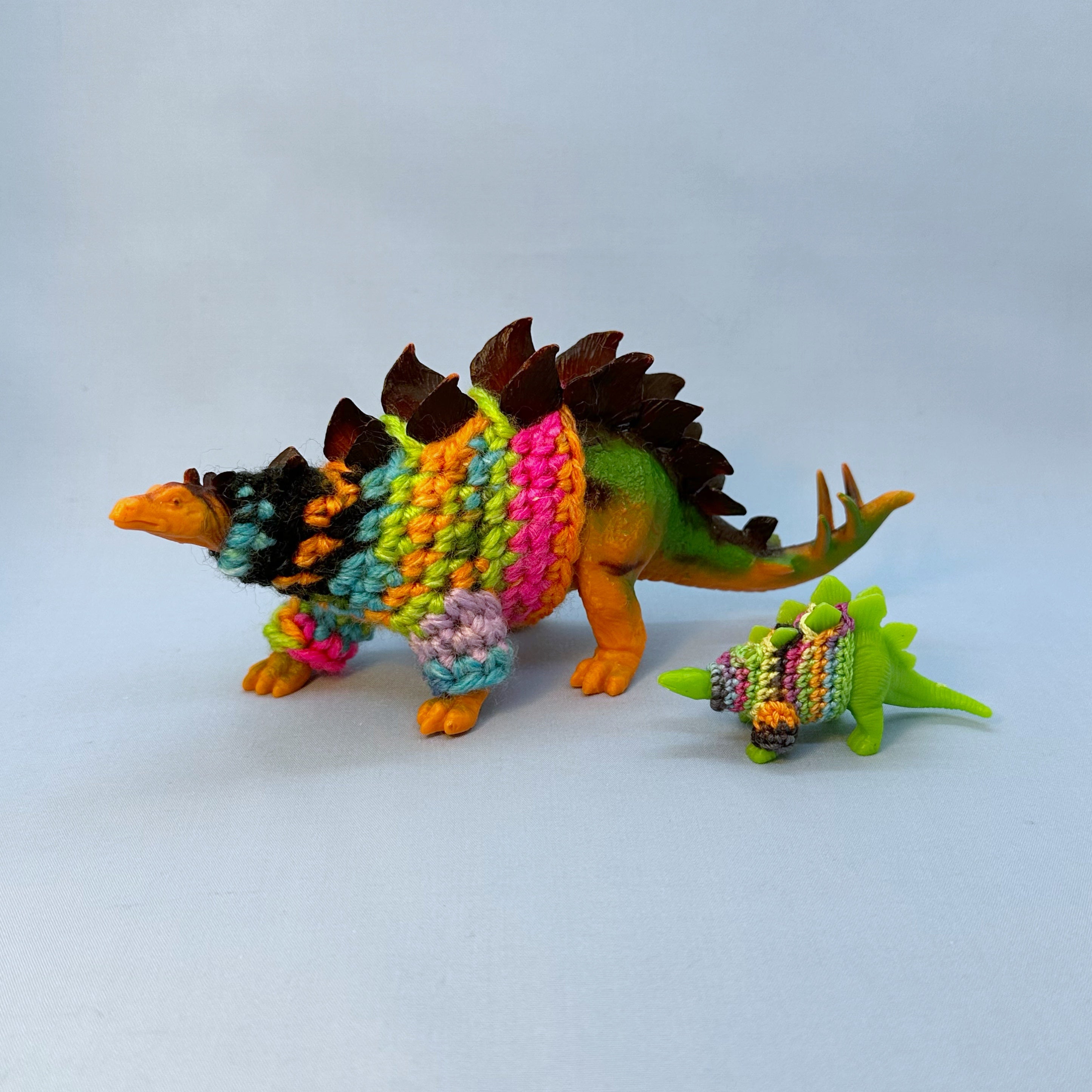 A large plastic toy stegosaurus wearing a hand crocheted multicolor sweater, with a smaller green toy stegosaurus also wearing a hand crocheted sweater. They stand against a blue background.